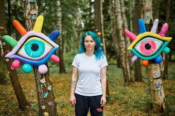 Weird young adult woman with dyed turquoise hair in white T shirt near big eyes art installation in...