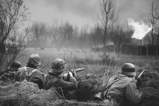 Re-enactors Armed Rifles And Dressed As World War Ii German Wehrmacht Infantry Soldiers Fighting Defensively In Trench. Defensive Position. Building On Fire On Background. Black And White Colors.