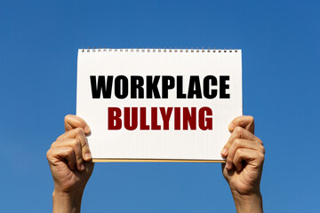 Workplace bullying text on notebook paper held by 2 hands with isolated blue sky background. This message can be used as business concept about workplace bullying.