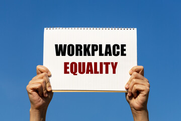 Workplace equality text on notebook paper held by 2 hands with isolated blue sky background. This message can be used as business concept about workplace equality.