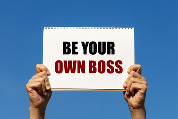 Be your own boss text on notebook paper held by 2 hands with isolated blue sky background. This...