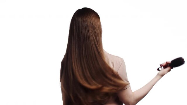 Woman in a pink t-shirt slowly combing her long healthy shiny brown hair with a comb in a back view on a white background. Woman getting her hair done. Hair care concept