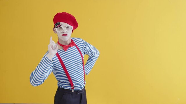 Portrait of a mime playing a funny performance, standing on a yellow background.The young mime guy puts his hands on his hips and shakes his finger in displeasure