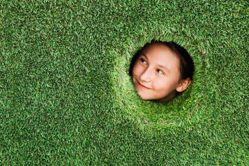 Artificial turf green background. Little kid looks into round hole in a wall made of synthetic turf...