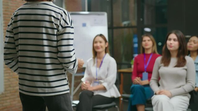 From a rear view, the female coach engaged with her audience in the meeting room, sharing her experiences and insights with a confident and assured demeanor.