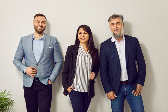 Small business team at work. Group portrait of three young and mature people in office. Two men and one woman in smart casual clothes standing by white office wall, looking at camera, and smiling