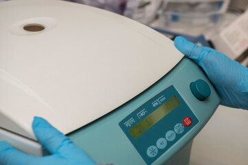 Two hands with blue nitril gloves next to the control panel of a closed centrifuge in a laboratory.