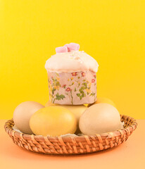 Easter cake and eggs in a wicker basket on a yellow background. Easter holiday. A bright Easter composition. Vertical photo. Close-up. Selective focus.