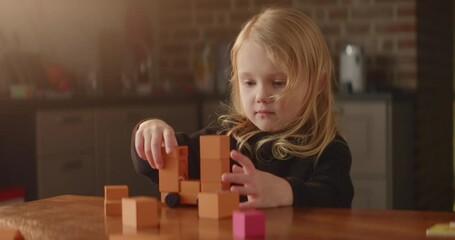 Blond girl plays busily on a table with orange wooden toy blocks, game with cubes, cute child builds a tower with wheels. Kids leisure activity. Active imagination. Nice home interior. HQ 4K video