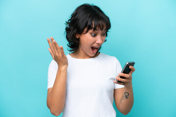 Young Argentinian woman isolated on blue background looking at the camera while using the mobile with surprised expression