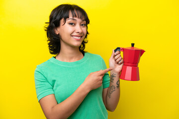 Young Argentinian woman holding coffee pot isolated on yellow background and pointing it