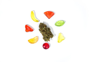 A dry bud of marijuana lies on a white background surrounded by chewy gummy candies of various...