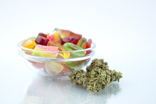 Assorted different gummy candies in a small glass bowl, near in the foreground several buds of dried medical marijuana, reflected on the glass