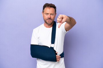 Middle age caucasian man with broken arm and wearing a sling isolated on purple background showing thumb down with negative expression