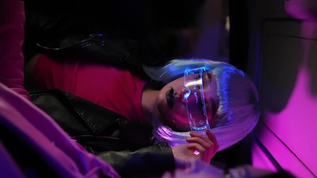 Portrait beautiful trendy woman wear white outfit and futuristic glasses posing looking into camera standing by illuminated wall in night club. Cyberpunk style, sensual look clubber in neon blue light