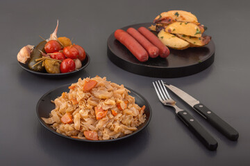 Stewed cabbage - bigos in a black plate against the background of pickled vegetables and baked potatoes with smoked sausages. Country style breakfast