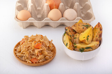 Braised cabbage - bigos and grilled sausage in a white porcelain plate against the background of chicken eggs and rustic potatoes.