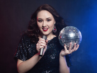 young woman wearing black dress, holding disco ball and singing into microphone over dark background