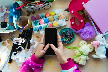 Hand of child girl using smartphone on chaotic desk in home office. Desk with objects for creativity, paints for drawing, children toys randomly scattered