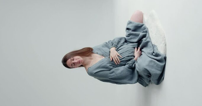 beautiful pregnant girl with blond long hair in a long dress sits on the floor on a white background waving her head that everything is fine and showing the ok sign with her hand. vertical video