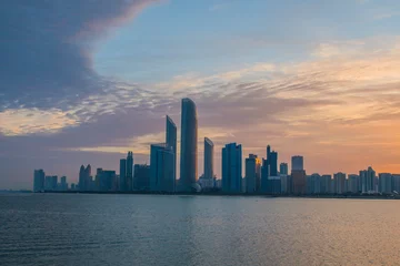 Schilderijen op glas Abu Dhabi cityscape at sunrise with clouds and water in front. © ElMehdi