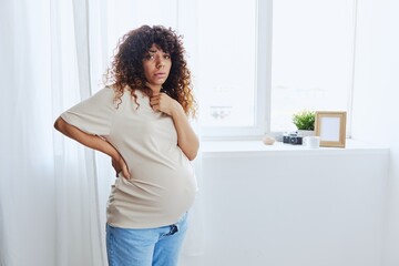 A pregnant woman stands in the house at the window, tired and has a headache, tears in a home T-shirt, the complexity of motherhood