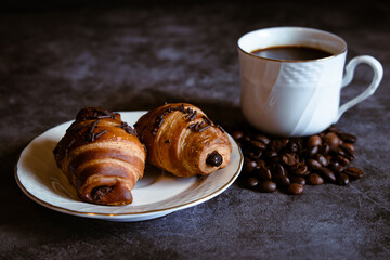 Cup of coffee with some chocolate croissants and coffee beans on a grayish background.