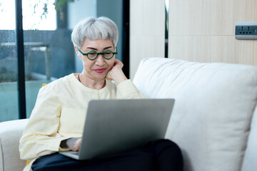 happy asian senior woman grey hair wear glasses using laptop computer sitting on couch in living room. relaxing elderly female having video chat. technology and old age concept.