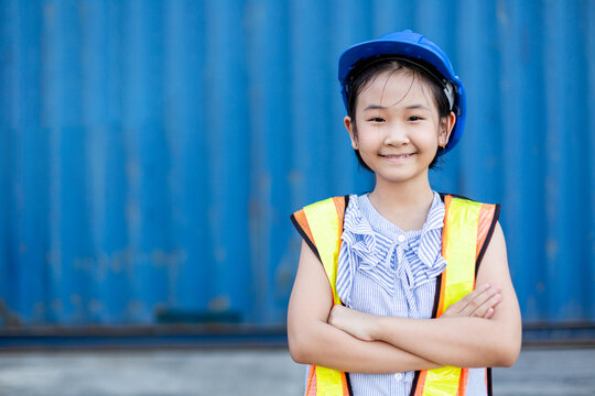 Happy smiling Asian little girl dress up engineer suit wearing hard hat crossed arms standing at containers port, Area logistics import export and shipping. future learning and inspiration concept