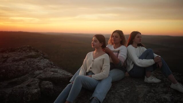 A mother and her daughters sit on top of a cliff during an incredible sunset. A woman braids her teenage daughter's hair. Happy family on vacation. A calm summer evening in nature