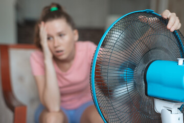 Young woman using electric fan at home in living room, sitting on couch cooling off during hot weather, suffering from heat, high temperature - obrazy, fototapety, plakaty