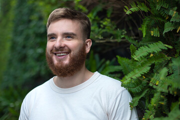 Naklejka na ściany i meble Portrait of happy young bearded man with beard breathing deep, inhale fresh air on natural background, green wall with leaves, flower in tropical country in garden. Eco, ecological concept. 