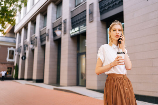 Attractive young woman in casual clothes holding takeaway coffee cup and talking on mobile phone during walking on city street in sunny summer day, looking away. Pretty blond female teenager outdoors. - Powered by Adobe