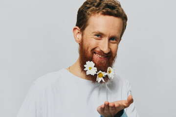 Portrait of a funny man in a white T-shirt with flowers daisies in his beard on a white isolated background, copy place. Holiday concept and congratulations.