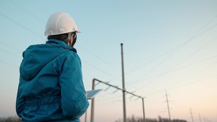 A woman power engineer in white helmet inspects power line using data from electrical sensors on a tablet. High voltage electrical lines at sunset. Distribution and supply of electricity. clean energy