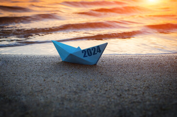 Blue paper boat with inscription 2024, on sandy shore beach against sunset.