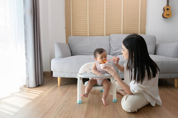 Young mother helping baby eating blend food on baby chair - obrazy, fototapety, plakaty