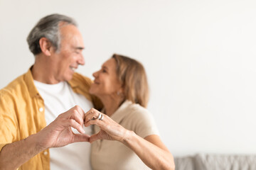 Loving senior couple embracing while doing heart shape gesture with hands, looking at each other and smiling, free space
