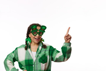 Beautiful woman wearing stylish carnival goggles with clover leaves, for St Patrick's pub party, pointing index finger at copy space, smiling looking at camera over isolated white background. Ad space