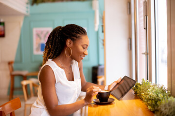 Young black woman drinking coffee while looking at digital tablet in the cafe