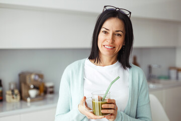 Portrait of smiling adorable middle-aged lady in blue cardigan holding glass of fresh vegetable...