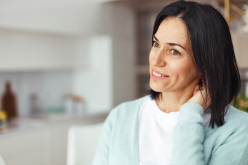 Portrait of shy charming mature brunette lady in blue cardigan standing in blurred home interior looking aside on copy space with smile, touching neck, feeling embarrassed and in love