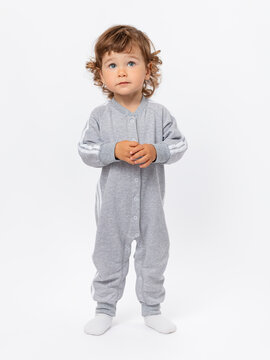 A 1-2-year-old toddler with curly hair stands in a gray jumpsuit and looks expressively up, fingering on a white background.