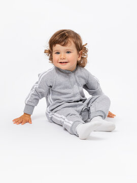 A 2-year-old toddler boy is sitting on the floor leaning on his hands and looking with interest with curly hair, plump cheeks in a gray jumpsuit and socks on a white background.