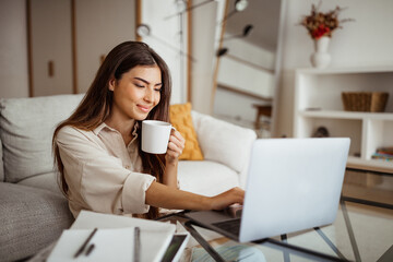 Cheerful young asian woman typing on computer and drinking coffee cup in room interior
