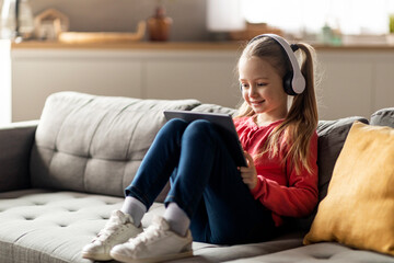 Cute Little Girl Wearing Wireless Headphones Using Digital Tablet At Home
