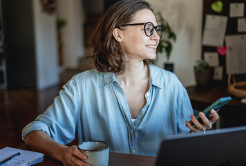 Young smiling adult woman with coffee mug looking at smartphone screen at workplace with laptop at...