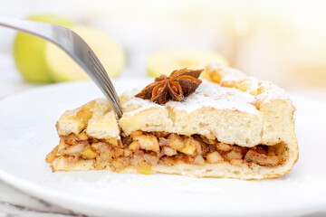 Piece of cinnamon apple pie sprinkled with powdered sugar and decorated with anise star on white plate. Hand breaks off piece of cake with a fork close-up. Delicious dessert in a cafe on a sunny day.