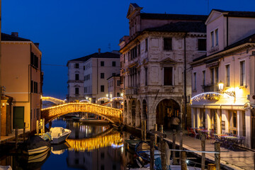 Chioggia cityscape in Venice Lagoon with narrow Vena water canal with colorful colorful boats among old buildings and illuminated brick bridge, blue evening sky, Veneto region, northern Italy