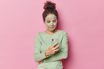 Fototapeta na wymiar Young beautiful woman with dark curly hair gathered in bun applies nourishing sheet mask takes care of facial skin undergoes beauty treatments wears casual green jumper holds mobile phone chats online